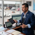 En su oficina, un director de operaciones de rasgos mexicanos y mediana edad revisa los flujos de trabajo. Viste un saco azul sobre una camisa clara y sostiene una tableta con una mano, mientras que con la otra señala un monitor que despliega gráficos y métricas de logística. Detrás de él, un gran ventanal revela el movimiento constante de un patio de carga lleno de camiones y contenedores. Su escritorio refleja el día a día de la operación: mapas detallados de la República Mexicana, reportes impresos y una computadora portátil abierta.